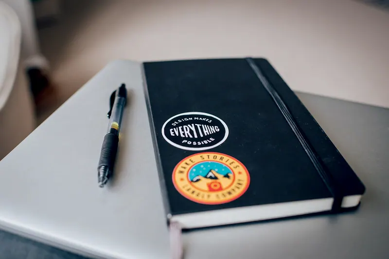Imagem ilustrativa: adesivos colados num caderno preto, ao lado de uma caneta